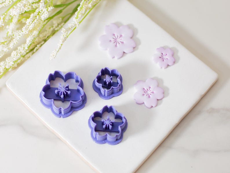 May include: Five purple plastic flower-shaped cookie cutters of varying sizes, with three pink fondant flowers on a white surface.