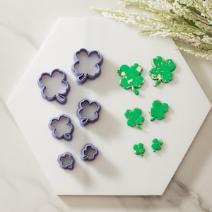 May include: A set of eight cookie cutters in the shape of four-leaf clovers. The cutters are purple and come in four different sizes. There are also four green four-leaf clovers on the white surface.