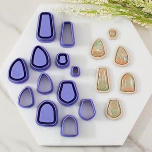 May include: A collection of polymer clay cutters in various shapes, including teardrops and rectangles, in a vibrant purple color. Next to the cutters are several finished clay pieces with a marbled effect in shades of green, orange, and beige. The items are arranged on a white hexagonal surface.