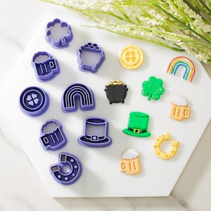 May include: A set of 14 purple and green plastic cookie cutters shaped like shamrocks, rainbows, beer mugs, gold coins, a pot of gold, a horseshoe, and a leprechaun hat.