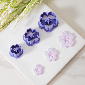 May include: Set of four purple plastic flower cookie cutters and four pink fondant flower shapes. The cutters are in descending size order, with the largest cutter at the top of the image. The fondant shapes are arranged in a similar order, with the largest shape at the top of the image.