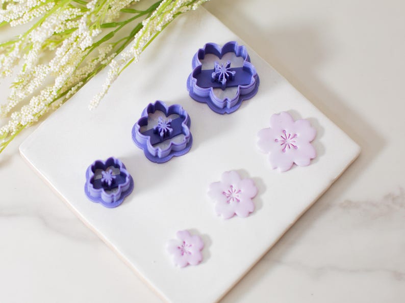 May include: Set of four purple plastic flower cookie cutters and four pink fondant flower shapes. The cutters are in descending size order, with the largest cutter at the top of the image. The fondant shapes are arranged in a similar order, with the largest shape at the top of the image.