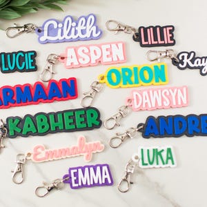 May include: A collection of personalized keychains featuring various names including Lilith, Lillie, Lucie, Aspen, Kayden, Armaan, Orion, Dawsyn, Kabheer, Andres, Emmalyn, Luka, and Emma. Each keychain has a metal lobster clasp and colorful lettering.