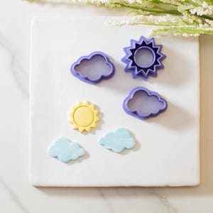 May include: Purple plastic cookie cutters in the shapes of a sun and two clouds. There are also two yellow and blue fondant cookies in the shapes of a sun and a cloud.