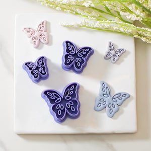 May include: Five purple and pink butterfly-shaped cookie cutters on a white surface. The cutters are different sizes and have intricate designs.
