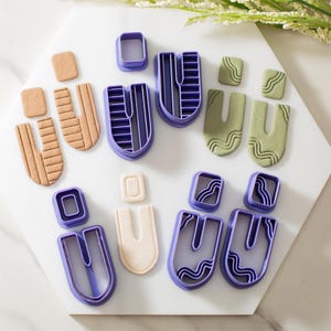 May include: A set of nine purple polymer clay earring cutters in various geometric shapes, including a rectangle, a square, and a U-shape. The cutters are arranged on a white surface.