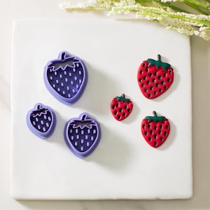 May include: A set of four cookie cutters in the shape of strawberries. Two cutters are purple and two are red. The cutters are arranged on a white surface.