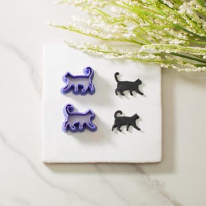 May include: Two purple plastic cat cookie cutters and two black fondant cat shapes on a white surface.