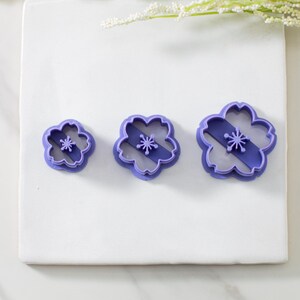 May include: Three purple plastic cookie cutters shaped like cherry blossoms. The cutters are different sizes, with the smallest cutter on the left and the largest cutter on the right.