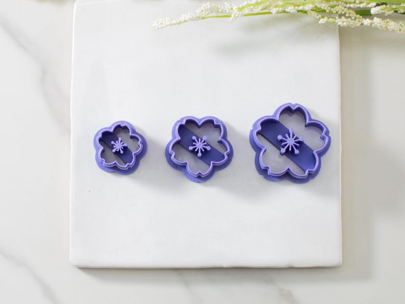 May include: Three purple plastic cookie cutters shaped like cherry blossoms. The cutters are different sizes, with the smallest cutter on the left and the largest cutter on the right.