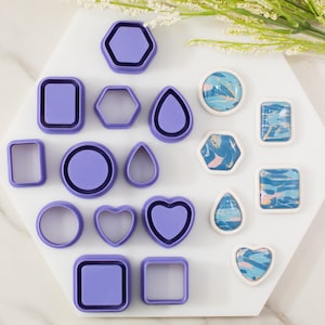 May include: A collection of purple polymer clay cutters in various shapes, including squares, circles, hexagons, and teardrops. Some cutters have matching polymer clay pieces with blue, pink, and white abstract designs. The items are arranged on a white hexagonal surface.