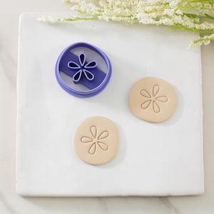 May include: A purple flower-shaped cookie cutter next to two beige cookies with flower imprints. The cookie cutter is round with a flower-shaped cutout. The cookies are round with a flower design.
