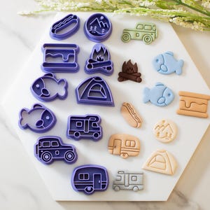 May include: A set of 16 purple plastic cookie cutters shaped like camping and outdoor-themed items, including a tent, campfire, fish, hot dog, RV, and car. The cutters are arranged on a white hexagonal surface.