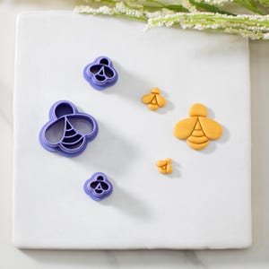 May include: A collection of bee-shaped cookie cutters in various sizes and designs. The cutters are made of purple plastic and yellow clay. The bee shapes include detailed wings and body segments, perfect for baking or crafting projects.