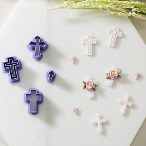 May include: A set of purple and white cross-shaped cookie cutters and fondant decorations. The cutters are in various sizes and styles, including a simple cross, a cross with a floral design, and a cross with a raised center. The fondant decorations are in white and feature a cross with a pink flower.