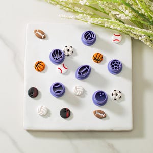 May include: A set of 14 purple plastic cookie cutters shaped like different sports balls, including a football, basketball, baseball, soccer ball, volleyball, and bowling ball. The cutters are arranged on a white surface.