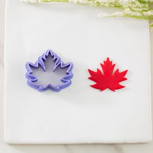 May include: A purple maple leaf-shaped cookie cutter next to a red maple leaf cutout. The cookie cutter is made of plastic and has a detailed outline. The red leaf is a solid color and appears to be made of paper or cardstock.
