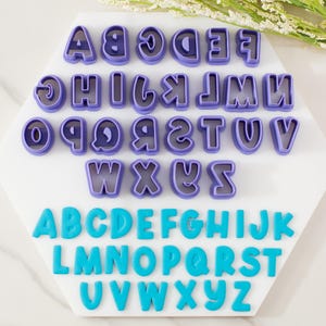 May include: A collection of purple letter-shaped cookie cutters and matching blue letters. The cutters are arranged on a white surface in a hexagonal shape. The letters are in a bold, rounded font, perfect for baking or crafting.