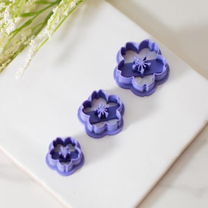 May include: Three purple plastic flower-shaped cookie cutters of different sizes. The cutters are on a white surface.