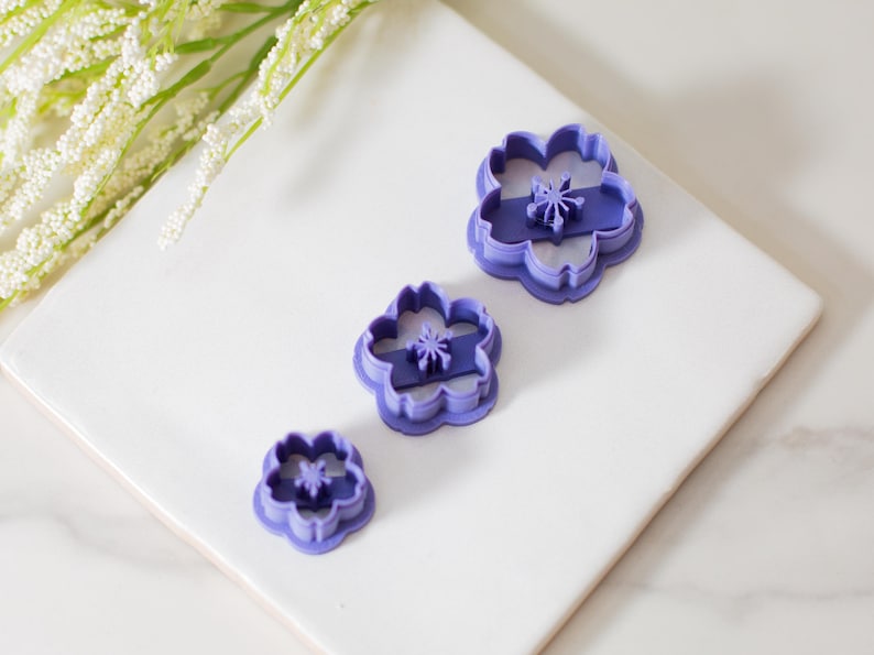 May include: Three purple plastic flower-shaped cookie cutters of different sizes. The cutters are on a white surface.