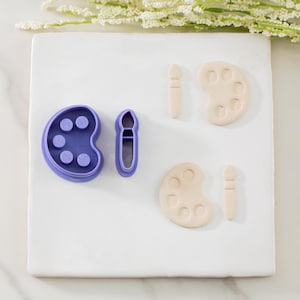 May include: A collection of art-themed cookie cutters. The set includes a purple palette and brush, along with two beige palettes and brushes. The items are arranged on a white surface, with a sprig of white flowers in the background.