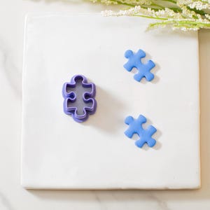 May include: Three purple plastic puzzle piece cookie cutters and two blue fondant puzzle pieces on a white surface.