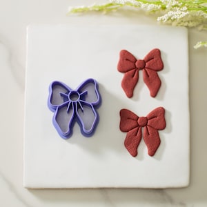 May include: A purple plastic bow cookie cutter with two red fondant bows on a white surface.
