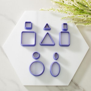 May include: A set of eight purple plastic cookie cutters in various shapes, including circles, ovals, squares, and triangles. The cutters are arranged on a white hexagonal surface.