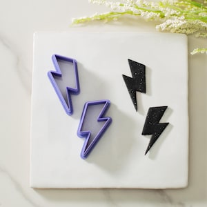 May include: Two purple plastic lightning bolt cookie cutters and two black fondant lightning bolts on a white surface.