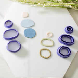 May include: A set of purple plastic cookie cutters in various shapes, including circles, ovals, and rounded rectangles. There are also three small pieces of clay in different colors: light blue, dark blue, and light green.