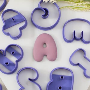 May include: A collection of purple plastic cookie cutters in various abstract shapes arranged around a central, smooth, dusty rose-colored letter A shaped cookie. The cutters feature open frames with internal structural bars for support.
