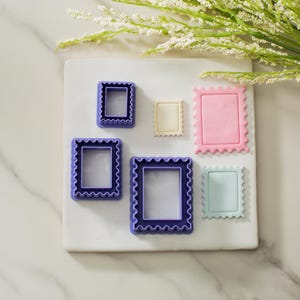 May include: A set of six purple and blue plastic cookie cutters shaped like different sized postage stamps. The cutters are on a white surface.