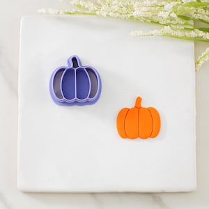 May include: Two pumpkin-shaped items on a white surface. One is a purple plastic cookie cutter, and the other is an orange pumpkin-shaped decoration. The background includes white flowers and greenery.