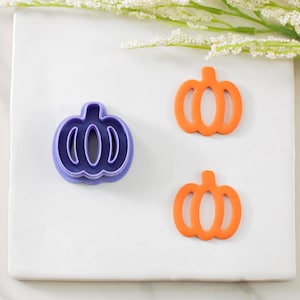 May include: Three pumpkin-shaped cookie cutters. One is purple, and the other two are orange. The cutters are on a white surface, with a sprig of white flowers in the background. Perfect for fall baking.