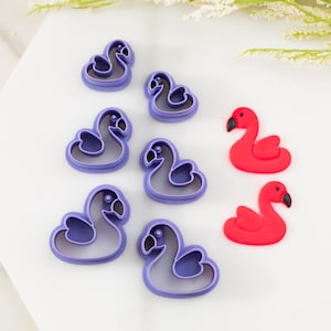 May include: An overhead shot shows flamingo-shaped cookie cutters and two red flamingo-shaped treats. The cookie cutters are purple with a white interior. The red flamingos have black beaks. The items are arranged on a white surface.