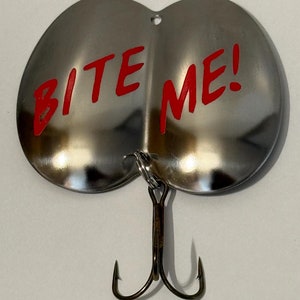 Puede incluir: Un se&ntilde;uelo de pesca plateado con dos anzuelos. El se&ntilde;uelo tiene forma de dos mitades redondeadas y tiene las palabras "BITE ME!" escritas en rojo en la superficie.