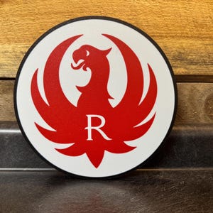Pu&ograve; includere: Un emblema rotondo bianco con un disegno di uccello rosso a forma di fenice e la lettera "R" al centro. L'emblema ha un bordo nero ed &egrave; posto su uno sfondo di legno.