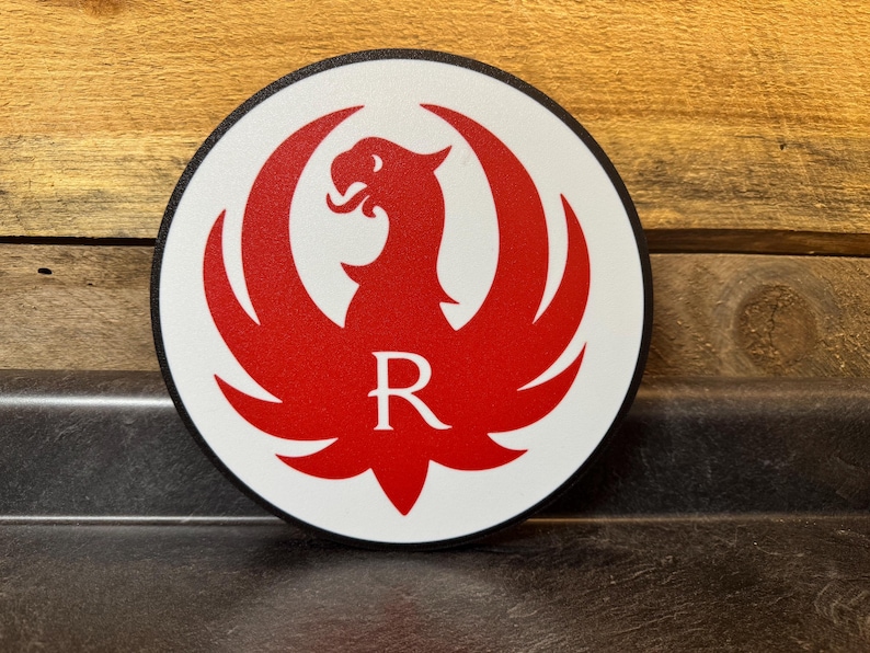 Pu&ograve; includere: Un emblema rotondo bianco con un disegno di uccello rosso a forma di fenice e la lettera "R" al centro. L'emblema ha un bordo nero ed &egrave; posto su uno sfondo di legno.
