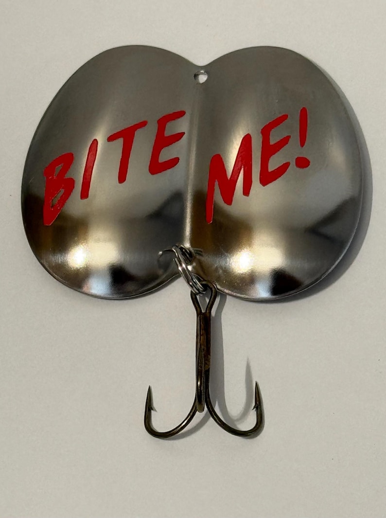 Puede incluir: Un se&ntilde;uelo de pesca plateado con dos anzuelos. El se&ntilde;uelo tiene forma de dos mitades redondeadas y tiene las palabras "BITE ME!" escritas en rojo en la superficie.