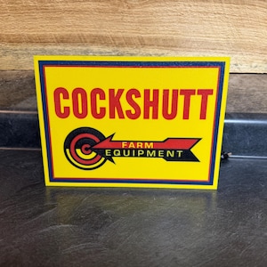 May include: Yellow sign with blue and black trim. The sign reads "Cockshutt Farm Equipment" with a red and black target graphic.