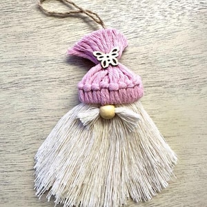 Może przedstawiać: Ręcznie robiona ozdoba gnoma z makramy. Gnom ma r&oacute;żową dzianinową czapkę z drewnianym motylem, białą brodę z frędzlami i drewnianą koralik. Do zawieszenia przymocowany jest sznurek jutowy. Ozdoba jest prezentowana na jasnej drewnianej powierzchni.