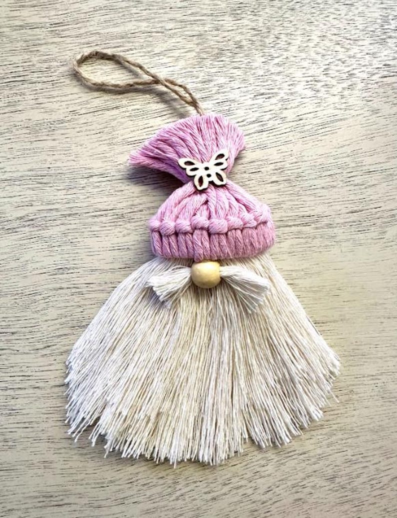 Może przedstawiać: Ręcznie robiona ozdoba gnoma z makramy. Gnom ma r&oacute;żową dzianinową czapkę z drewnianym motylem, białą brodę z frędzlami i drewnianą koralik. Do zawieszenia przymocowany jest sznurek jutowy. Ozdoba jest prezentowana na jasnej drewnianej powierzchni.