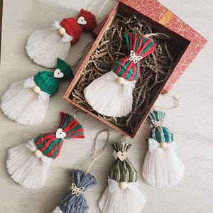 Może przedstawiać: Ręcznie robione ozdoby świąteczne w kształcie gnom&oacute;w z białymi brodami z przędzy, kolorowymi czapkami z dzianiny i nosami z drewnianych koralik&oacute;w. Ozdoby znajdują się w ozdobnym pudełku z papierowym wypełnieniem, idealne na święta.