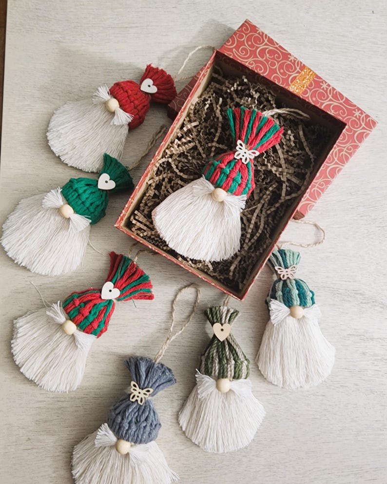 Może przedstawiać: Ręcznie robione ozdoby świąteczne w kształcie gnom&oacute;w z białymi brodami z przędzy, kolorowymi czapkami z dzianiny i nosami z drewnianych koralik&oacute;w. Ozdoby znajdują się w ozdobnym pudełku z papierowym wypełnieniem, idealne na święta.