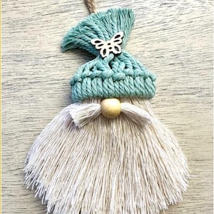 Może przedstawiać: Ręcznie robiona ozdoba gnoma z makramy z turkusową dzianinową czapką, drewnianym motylem i długą, kremową brodą. Drewniana koralik tworzy nos. Pętelka z juty jest przymocowana do zawieszenia.