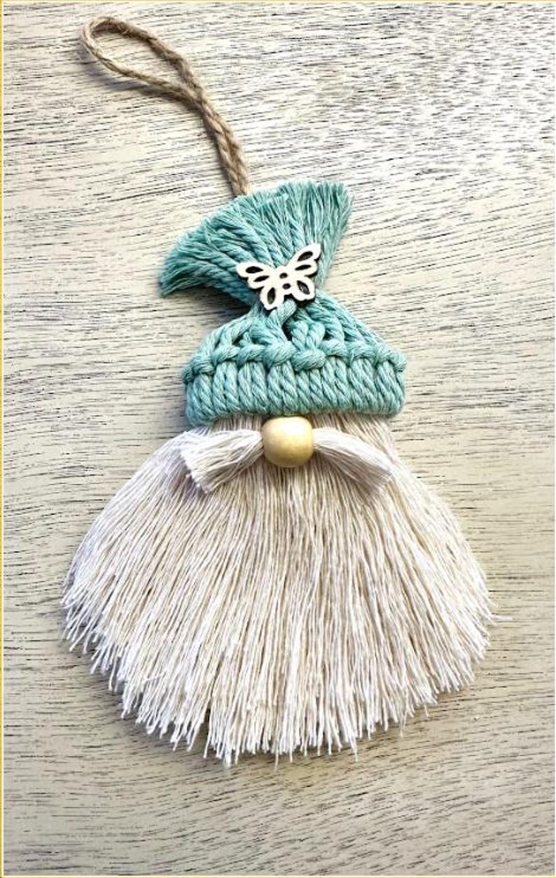 Może przedstawiać: Ręcznie robiona ozdoba gnoma z makramy z turkusową dzianinową czapką, drewnianym motylem i długą, kremową brodą. Drewniana koralik tworzy nos. Pętelka z juty jest przymocowana do zawieszenia.