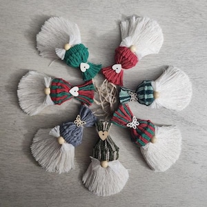 Może przedstawiać: Ręcznie robione ozdoby w kształcie gnom&oacute;w z makramy z białej, zielonej, czerwonej i niebieskiej przędzy. Każdy gnom ma białą brodę z frędzlami, drewniany nos z koralik&oacute;w i dekoracyjną kokardę lub czapkę. Idealne na dekoracje świąteczne.