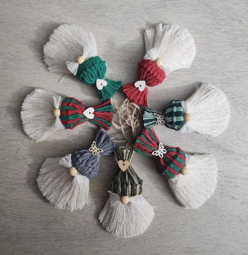 Może przedstawiać: Ręcznie robione ozdoby w kształcie gnom&oacute;w z makramy z białej, zielonej, czerwonej i niebieskiej przędzy. Każdy gnom ma białą brodę z frędzlami, drewniany nos z koralik&oacute;w i dekoracyjną kokardę lub czapkę. Idealne na dekoracje świąteczne.