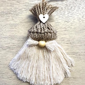 Może przedstawiać: Ręcznie robiona ozdoba gnoma z dzianinową szarą czapką, drewnianym sercem i białą brodą z frędzlami. Ozdoba ma pętlę z juty do zawieszenia i drewniany akcent z koralik&oacute;w. Tło to jasna drewniana powierzchnia.