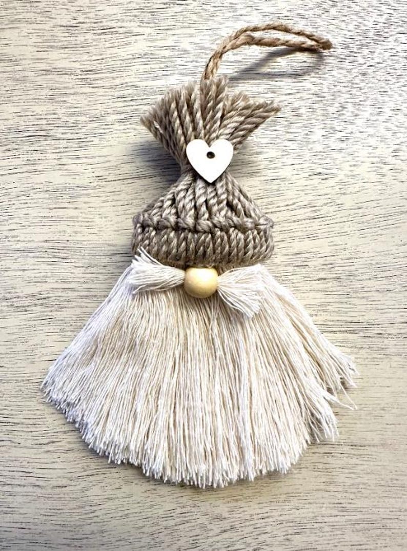 Może przedstawiać: Ręcznie robiona ozdoba gnoma z dzianinową szarą czapką, drewnianym sercem i białą brodą z frędzlami. Ozdoba ma pętlę z juty do zawieszenia i drewniany akcent z koralik&oacute;w. Tło to jasna drewniana powierzchnia.